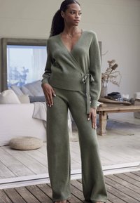 Pullover oversized verde oliva con design avvolgente e cintura, abbinato a pantaloni a gamba larga. Tessuto morbido, polsini a costine e scollatura a V.