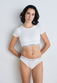 Žena s krátkými tmavými vlasy, oblečená v bílém crop topu od Calvina Kleina a odpovídajícím prádle, stojící na jednolitém bílém pozadí.