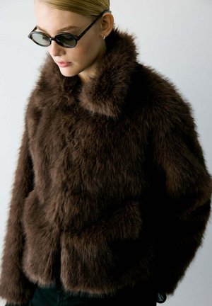 Femme portant une veste en fausse fourrure marron foncé et des lunettes de soleil ovales noires, regardant vers le bas avec les cheveux tirés en arrière sur un fond clair.