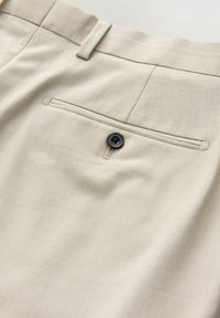 Pantalones beige con una tela texturizada, que presentan un bolsillo trasero con cierre de botón negro y un sutil detalle de costura.