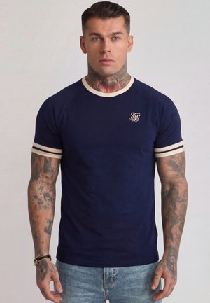 Mann mit Hals- und Arm-Tattoos, der ein marineblaues, eng anliegendes T-Shirt mit hellem Besatz an Kragen und Ärmeln trägt und vor einem einfarbigen Hintergrund steht.