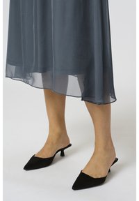 Mules en suede noir avec un bout pointu et un petit talon stiletto, portés avec une robe grise transparente et fluide qui a un ourlet en couches.