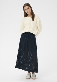 Pull crème en maille torsadée avec encolure ronde, associé à une longue jupe noire transparente à motif floral. Des chaussures décontractées complètent la tenue.