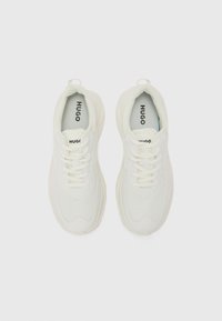 White sneakers with textured fabric, rounded toe, lace-up design, and "HUGO" branding on the tongue and insole. Soft, cushioned sole.