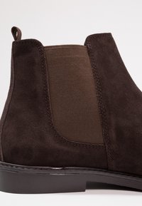 Botín de ante marrón que presenta paneles laterales de lona, una lengüeta en la parte superior, suela lisa y costuras a la vista, mostrando un diseño moderno y elegante.