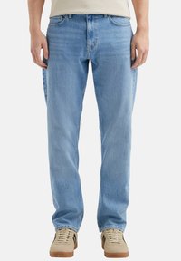 Jean en denim bleu clair avec une coupe décontractée, présentant un léger effet usé et un design classique à cinq poches. Porté avec des baskets beige.