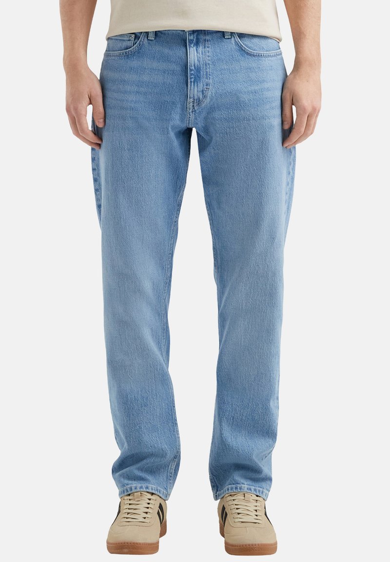 Jean en denim bleu clair avec une coupe décontractée, présentant un léger effet usé et un design classique à cinq poches. Porté avec des baskets beige.