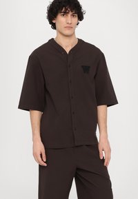 Brown short-sleeved button-up shirt with a henley neckline and a decorative W logo on the left chest. Soft fabric texture.