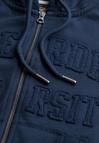 Hoodie zippé bleu marine avec le texte brodé "SUPERDRY UNIVERSITY", fermeture éclair en métal, cordons avec embouts en métal et texture de tissu douce.