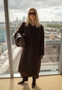 Długi brązowy płaszcz z teksturowanej tkaniny, nałożony na ciemny strój. Dodatki to duże okulary przeciwsłoneczne i pleciona brązowa torba.