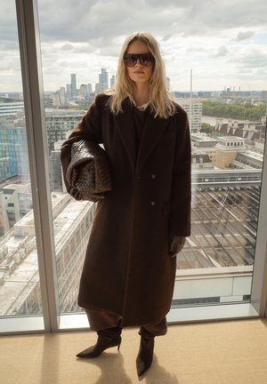 Ilgas rudas paltas, pagamintas iš tekstūruoto audinio, uždedamas ant tamsios aprangos. Aksesuarai apima didelius saulės akinius ir nertą rudą rankinę.