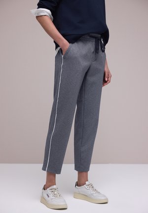 Person iført grå joggebukser med hvid sidestrejf, navy top og hvide sneakers, stående med den ene hånd i lommen.