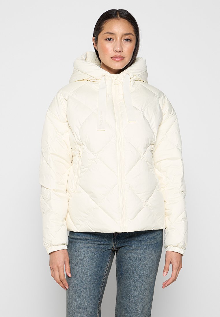 Gequilted witte pufferjack, ritssluiting, verstelbare capuchon, voorzien van diagonale stikpatronen en zijzakken, gecombineerd met spijkerbroeken.