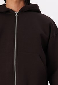 Hoodie zippé marron avec une poche avant, texture douce et capuche. Comprend une fermeture éclair argentée et un design sans coutures pour un ajustement confortable.