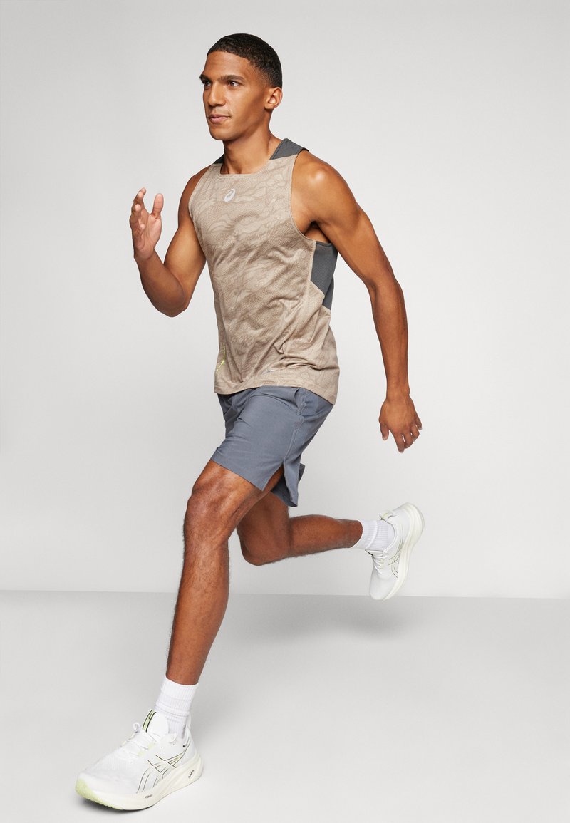 Top sin mangas marrón claro con patrón texturizado y espalda gris oscuro contrastante, combinado con pantalones cortos grises y zapatillas deportivas blancas con detalles amarillos.