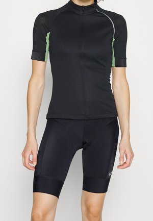 Frau trägt ein schwarzes Kurzarm-Radtrikot mit grünen Seitenpaneelen und schwarze gepolsterte Radhosen vor neutralem Hintergrund.