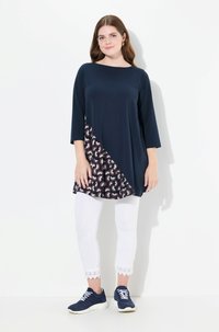 Marinblå tunika med trekvartsärmar, med en mönstrad asymmetrisk nederkant i burgundy och krämvit; matchad med vita leggings och marinblå sneakers.