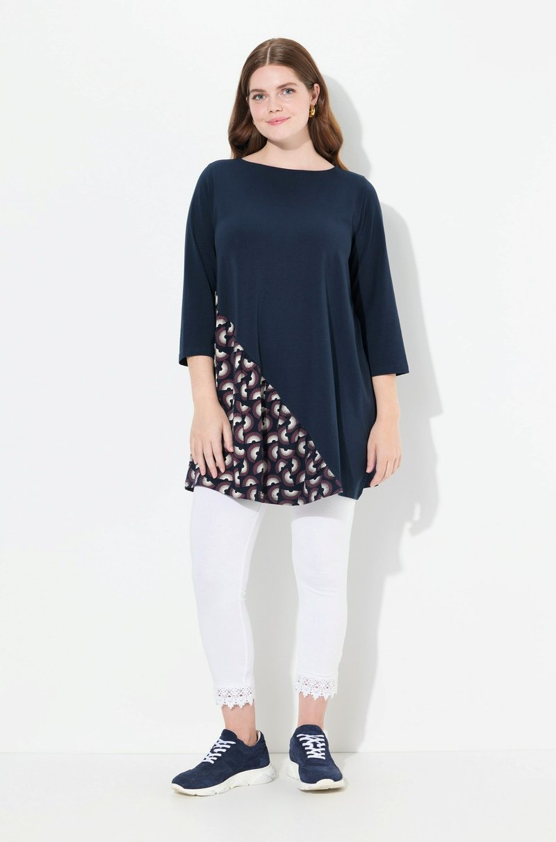 Marinblå tunika med trekvartsärmar, med en mönstrad asymmetrisk nederkant i burgundy och krämvit; matchad med vita leggings och marinblå sneakers.
