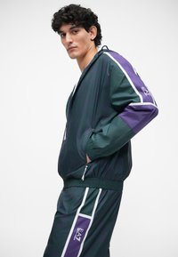 Trainingsjacke und Hose in Dunkelgrün mit lila und weißen Akzenten, ausgestattet mit einem Reißverschluss vorne, elastischen Bündchen und einem Logodetail am Ärmel.