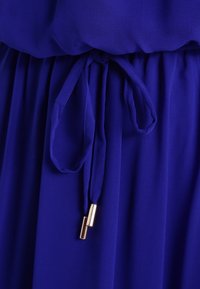 Livlig kunglig blå klänning med lätt material, samlad midja och en knytning med metalliska guldaccenter. Slät textur.