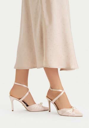 Frau trägt hellrosa Satin-High-Heels mit Knöchelriemen und Perlenverzierungen, kombiniert mit einem beigen Rock mit kleinen Punkten.