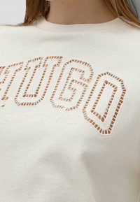 Sudadera color crema con letras recortadas y acentos cosidos. Textura suave, cuello redondo y detalle decorativo de bordado.