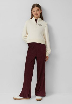 Felpa crema con zip e logo, abbinata a pantaloni larghi bordeaux in tessuto; sneakers bianche con suola beige.