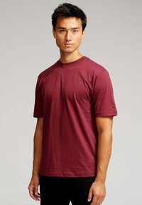 Ung mand med kort mørkt hår iført en ensfarvet mørkerød t-shirt med korte ærmer, der står foran en lys grå baggrund.