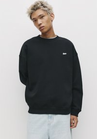 Sort sweatshirt lavet af cotton med rund hals og ribstrikkede manchetter, med et lille hvidt logo på venstre bryst. Bæret sammen med lyse jeans.