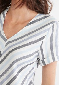 Raidallinen kangastoppi, jossa on V-aukko ja vaaleansinisiä, tummansinisiä sekä valkoisia vaakaraitoja. Lyhyet käärityt hihat ja pehmeä rakenne.