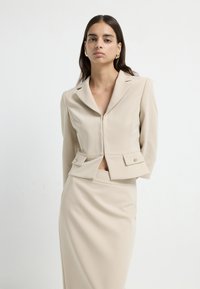 Beige dvoudílný outfit, který zahrnuje dobře padnoucí blazer se zipem, klopami s výstřihem do tvaru V a dvěma předními kapsami s klopou. Sukně má hladkou strukturu.