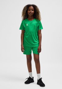 Grön sportuniform av lätt material, med kortärmad topp och shorts, kombinerad med svarta träningsskor och vita strumpor.