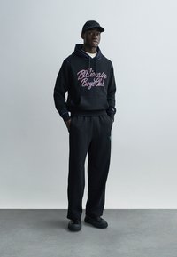 Μαύρο φούτερ με κουκούλα με το λογότυπο "Billionaire Boys Club" σε ροζ και μωβ, συνδυασμένο με μαύρο φόρμας και μαύρα αθλητικά παπούτσια. Η στολή έχει άνετη εφαρμογή.