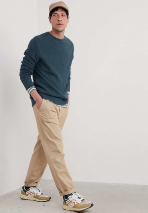 Pull à mailles texturées teal superposé sur une chemise blanche à col, associé à un pantalon beige et des baskets multicolores. Tenue décontractée et moderne.