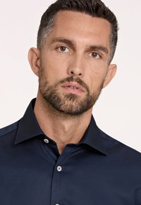 Camisa azul marino hecha de tela suave, con un collar clásico y botones. Primer plano del área del cuello, destacando la textura de la piel y la barba.