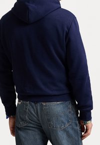 Man som bär en mörkblå hoodie och slitna blå jeans, sedd bakifrån med avslappnade händer vid sidorna.