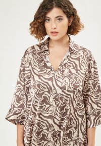 Abstract patroon shirt in bruin en crèmekleurige tinten, met een knoopsluiting aan de voorkant, kraag en korte mouwen. Materiaal lijkt glad en lichtgewicht.