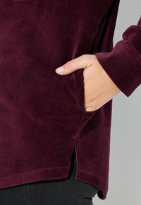 Burgundská kordová blůza s boční kapsou, zaobleným lemem a texturovanou tkaninou. Rukávy mají přiléhavé a mírně prodloužené manžety.