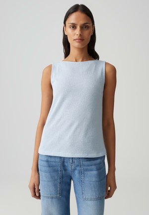 Femme aux longs cheveux foncés portant un haut sans manches bleu clair et un jean bleu à poches plaquées, sur un fond clair uni.