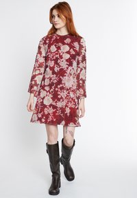 Rotes Blumen Kleid mit langen, leicht ausgestellten Ärmeln, einem runden Ausschnitt und einer fließenden Silhouette. Kombiniert mit kniehohen braunen Lederstiefeln.
