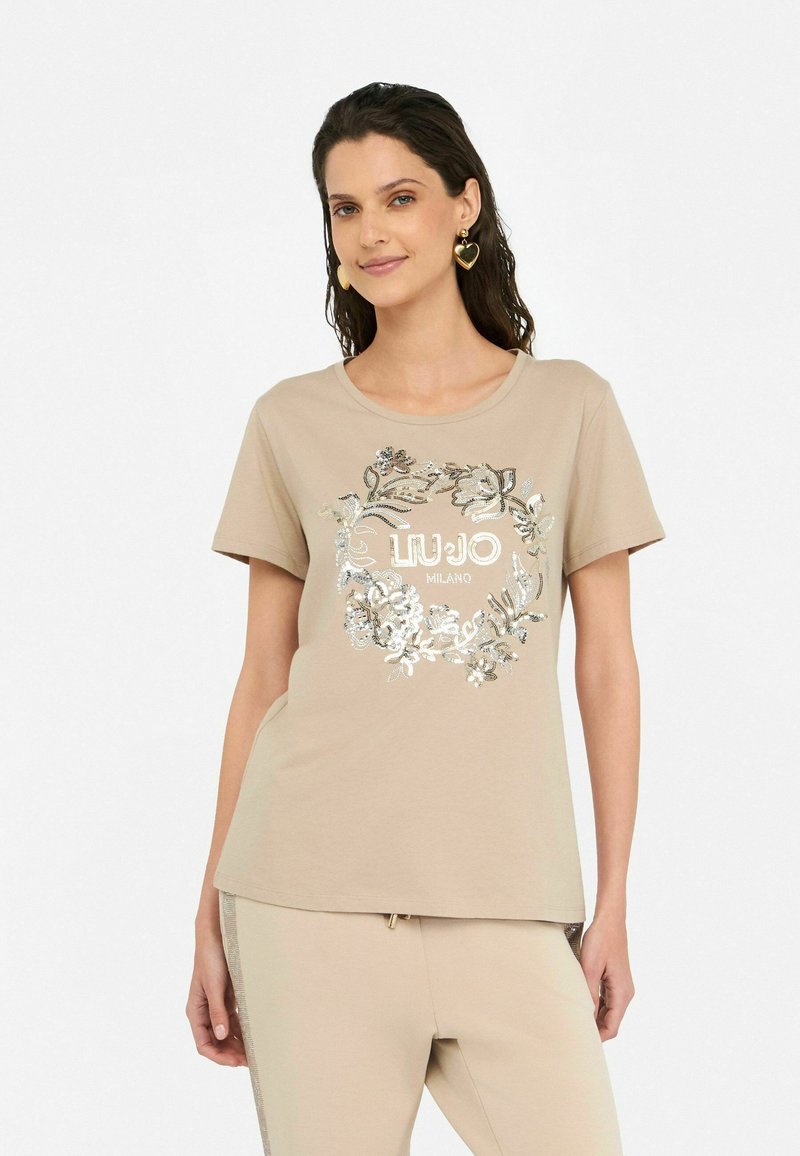 T-shirt beige à manches courtes avec un col rond, mettant en avant un design floral en sequins et le texte « El Lujó Milano » en argent. Matériau en coton doux.
