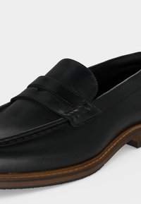 Mocassin en cuir noir avec une bride cousue sur le dessus, semelle marron avec coutures visibles et petit talon en bois.