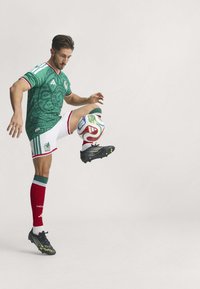 Green football jersey with a graphic pattern, white shorts, red knee-high socks, and black cleats. Player balances a colorful ball on one foot.