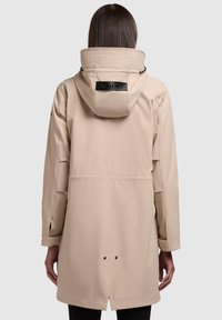 Veste imperméable beige avec un col haut, un détail de logo noir au dos, une coupe décontractée et des poches fendues sur les côtés. Texture lisse et mate.