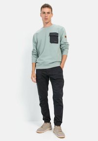 Lys grøn sweatshirt med en sort brystlomme, parret med sorte bukser og beige sneakers. Blødt stof, afslappet pasform og praktisk design.