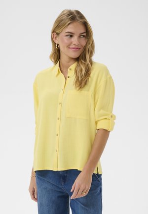 Mosolygó nő hullámos hajjal, laza sárga gombos ingben, redőzött ujjúval és kék farmerral, egy egyszínű háttér előtt állva.