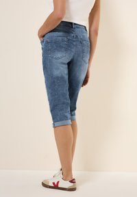 Pantalones capri de mezclilla en azul desvaído, con dobladillos enrollados, cintura de altura media y bolsillos traseros cosidos. Combinados con zapatillas blancas.