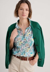 Cardigan verde tricotat peste o cămașă cu model care prezintă designuri florale colorate și abstracte; pantaloni bej cu talie înaltă și curea maro.