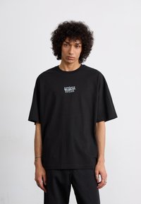 Sort oversized t-shirt lavet af bomuld, med et hvidt logo i gotisk stil på brystet og korte ærmer. Ensfarvet bag, glat tekstur.