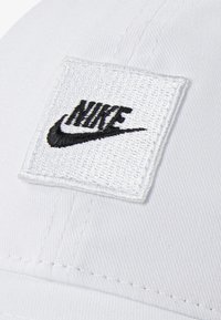 Căciulă de baseball albă, cu un material texturat, având un patch brodat cu un logo pătrat, care afișează "NIKE" în text negru și un swoosh negru.